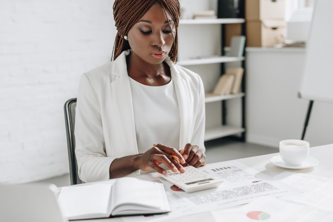focused african american adult businesswoman in white formal wear working at office desk and using calculator